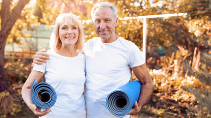 A happy senior couple stands smiling while holding their yoga mats in a bright and sunny park. The trees have autumn leaves, and they look ready for their yoga session together.