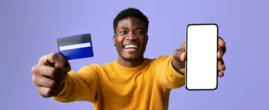 A man is happily displaying a credit card in one hand and a smartphone in the other. He is wearing a yellow sweater and has a joyful expression, indicating excitement about shopping.