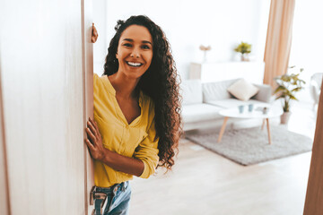 Fototapeta premium A woman with curly hair stands by the door, smiling warmly as she welcomes guests into her stylish living room. The space is bright with modern decor and comfortable furniture.