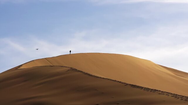 Traveler Slowly Climbs the Dune. A large sand dune is lit by the evening sun. In the distance, a man moves along its edge. He is accompanied by a quadrocopter