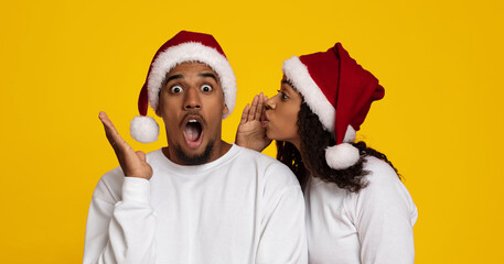 A man reacts with surprise while a woman whispers in his ear, both wearing festive Santa hats. The bright yellow background adds to the joyful holiday atmosphere.