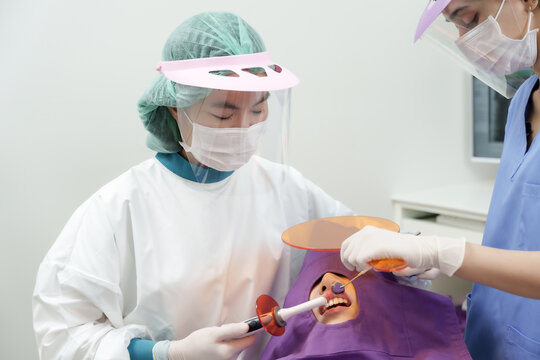 Asianfemaledentist wearing face shield and mask using curing light tool assisted by dental assistant to perform tooth filling procedure on adult asian woman during dental treatment session