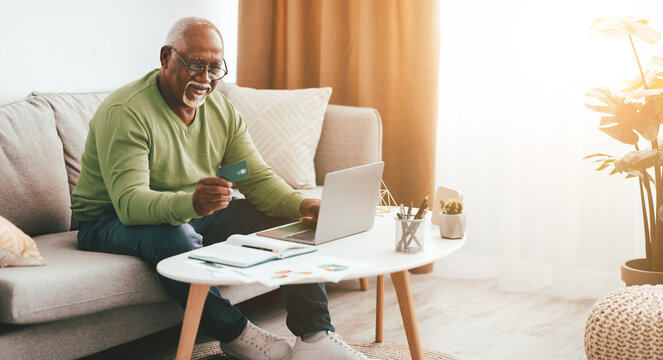 A senior man sits comfortably on a sofa, using a laptop to shop online. He holds a credit card in one hand, focused on the screen. Natural light brightens the room. - Powered by Adobe