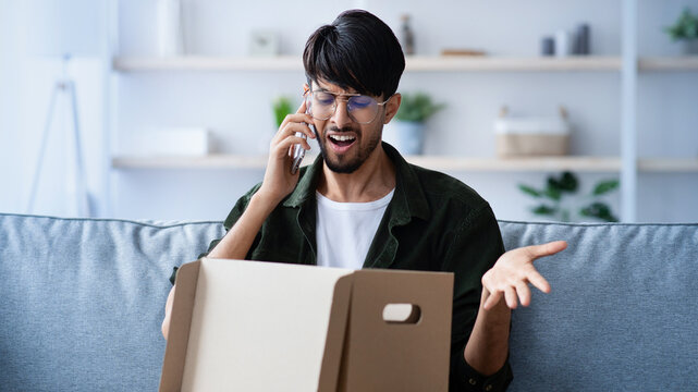 A man sits on a couch, looking surprised while talking on the phone. He opens a cardboard box, revealing unexpected items. The cozy living room features plants and light decor.