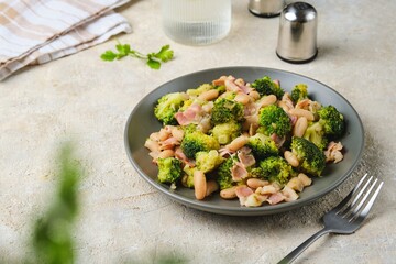 Fried broccoli with bacon and canned white beans on a gray plate against a light concrete background. Broccoli recipes.