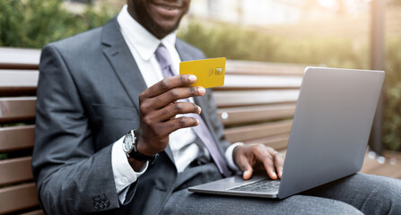 A man in a suit sits on a park bench, smiling as he holds a yellow credit card in one hand and...
