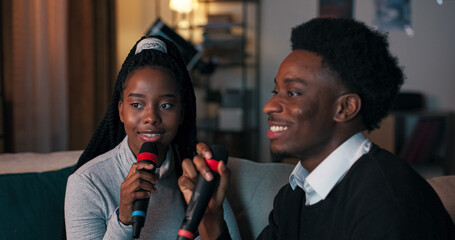 A close-up of two young friends of multi-racial background singing karaoke. The girl with dark hair wears a gray shirt, and the guy in a black sweater holds a microphone while smiling.