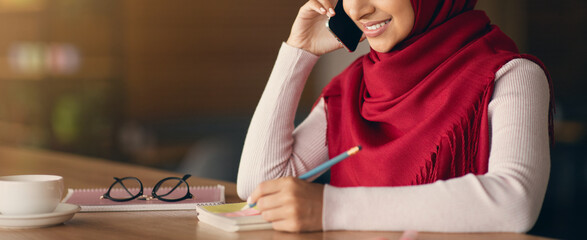 A woman wearing a red scarf talks on her phone while sitting at a wooden table in a cozy cafe. She writes notes in her notebook and has a cup of coffee beside her.