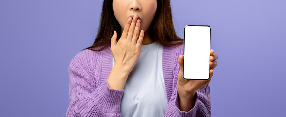A young woman stands against a soft purple background, expressing surprise as she covers her mouth and holds a blank smartphone. She wears a cozy sweater and looks directly at the viewer.