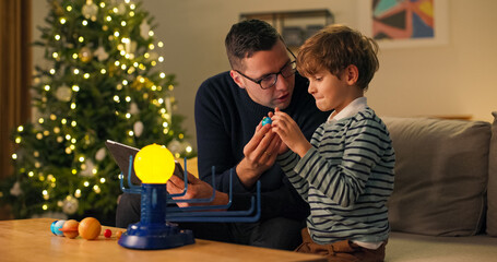 A child plays by a Christmas tree lit with lights, a school-age boy looks at a toy planetary system he built himself, fun combined with learning, the kid holds the planet.