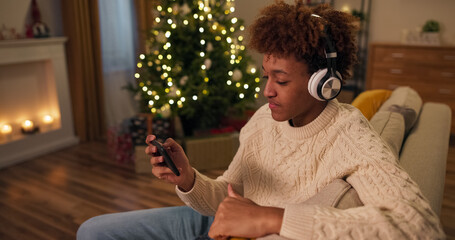 On a festive winter evening, a young boy sits on a couch by a Christmas tree lit with lights. He's...