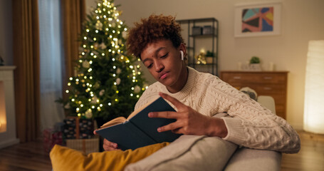 On a winter evening by the Christmas tree, a boy is listening to music on wireless headphones. He is reading an interesting book, turning the pages to immerse himself in the story.