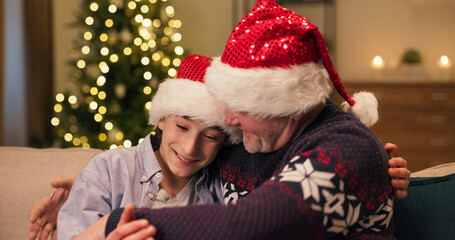 A close-up captures a father and son hugging tightly. They greet each other for the winter holidays, smiling with Santa hats on their heads. The young man wears a shirt, and the father wears a holiday