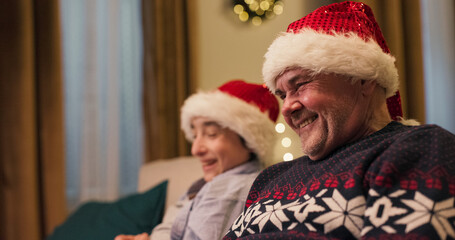 Close-up of middle-aged man and teenager sitting on sofa laughing out loud. They are wearing Santa hats. The man is dressed in warm holiday sweater. A Christmas tree flickers behind.