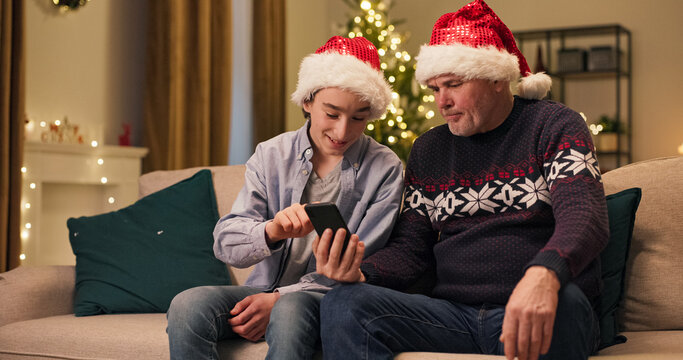 Sitting on a couch in the living room on Christmas Eve, a dad and son communicate. The attentive father holds the phone, and the son shows him the correct way to use a new program. Both wear Santa