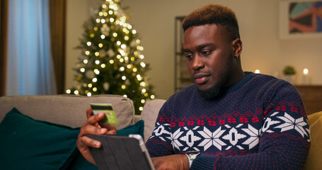 Holding a tablet and bank card, a young student of multi-racial background makes an order in an online store. He orders a gift for his girlfriend, and after the purchase is complete, he is happy and