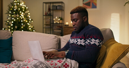 Covered with a blanket, a young man of multi-racial background sits on a sofa, typing on his laptop keyboard. He wears a warm sweater, and a Christmas tree is flickering nearby in the living room.