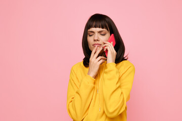 A woman in a yellow hoodie holds a pink phone, eyes closed and thoughtful, as she chats softly in a bright studio setting, creating a warm, friendly vibe.