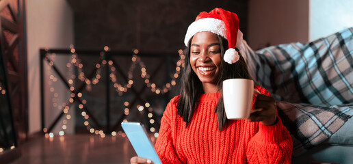 A woman wearing a bright red sweater and Santa hat sits comfortably on the floor with a mug in one hand and a tablet in the other, surrounded by holiday decorations and warm lights.