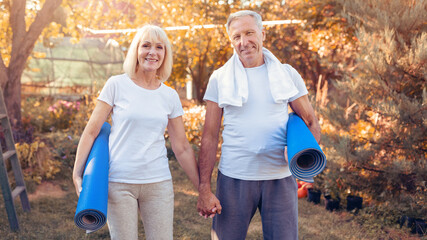 A happy senior couple stands in a sunny garden holding yoga mats. They are smiling and dressed in comfortable athletic clothing, ready to start their outdoor yoga session.