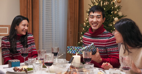 Sitting at the festive table, a girl with long black hair hands a gift to a boy. The man, wearing the same sweater as the girl, opens the gift, rejoices, looks at the camera, and smiles.