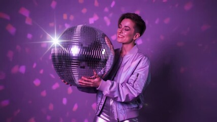 Cheerful person with a short haircut holding a large, shiny disco ball in a club setting, smiling at the camera under purple lights with falling confetti for a fun retro celebration - Powered by Adobe