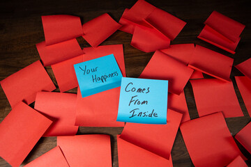 Blue sticky notes on a pile of red sticky notes on a dark wood surface with text reading Your Happiness Comes from Inside.