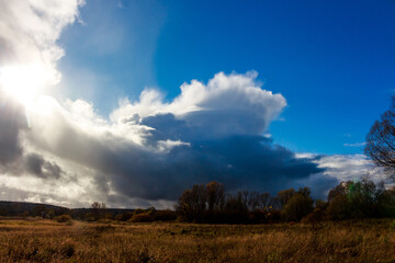 Dramatic cumulonimbus clouds fill a vibrant autumn sky. Bright sunrays pierce through dark shadows, illuminating a peaceful fall field