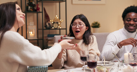 Friends sit at the festive table and give gifts. A brunette with long hair gives a gift to a multi-ethnic girl in glasses, who opens the gift and is satisfied. Both girls are happy and smiling.