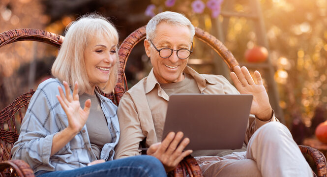 A happy couple sits in wicker chairs in a garden, enjoying a video call on a laptop. The warm sunlight creates a cozy atmosphere as they smile and wave, feeling connected. - Powered by Adobe