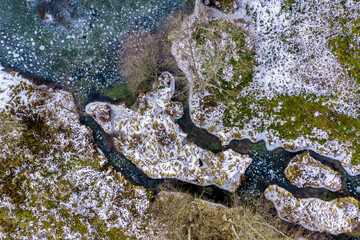 Top-down drone view of a meandering, snow-dusted stream with ice floes and mossy islands in the Black Forest, Germany