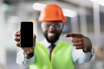 Smiling handsome african american young man in safety vest and helmet civil engineer or architect showing mobile phone with blank screen, mockup, using modern technologies, copy space