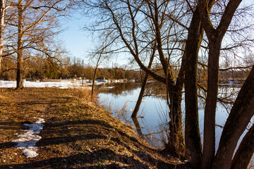 Obraz premium Spring thaw panorama: bare trees flank a river with melting ice and snow patches. Calm water reflects the clear sky, signaling nature's fresh start