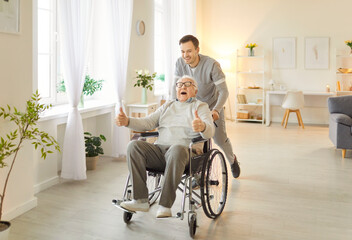 Joyful senior man with grey hair involved in rehabilitation procedure with young caregiver, using wheelchair for mobility as has walking , having fun in retirement house.