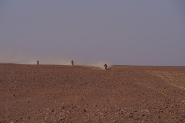 Three motorcyclists racing across a vast Algerian desert plain, kicking up dust in an off-road rally
