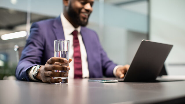 Glass of water in positive young black man manager, businessman drinking water while working on laptop at office, typing on computer keyboard, making marketing research or sending emails, copy space