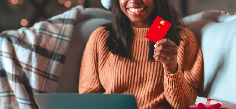 A woman with a joyful expression sits on a cozy couch, holding a red credit card in one hand and typing on a laptop. Holiday decorations are visible around her, creating a festive atmosphere.
