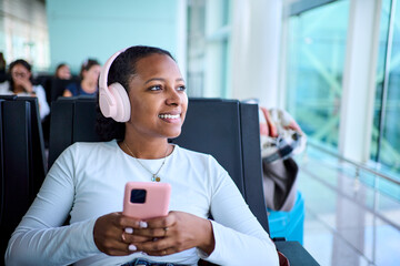 Smiling african woman with headphones sits in an airport lounge holding a smartphone, listening while waiting to board. Bright glass terminal and soft reflections convey a calm travel mood.