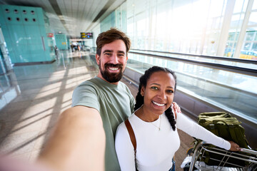 Happy interracial couple taking a selfie at an airport terminal, smiling beside a luggage trolley....