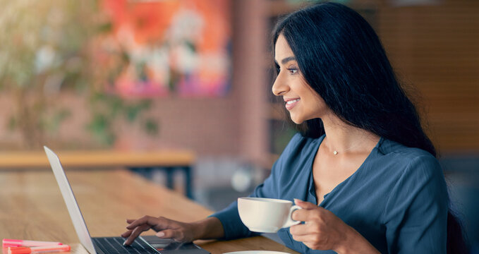 A young woman sits at a wooden table in a bright cafe. She smiles while typing on her laptop, holding a cup of coffee in her other hand. Colorful artwork decorates the background.