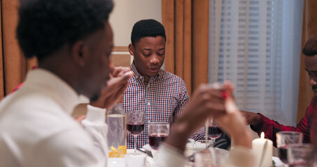 Following tradition, a group of young people sit at the table with closed eyes and held hands to pray before their dinner; the guy in the shirt says the prayer aloud, then they release hands, smile