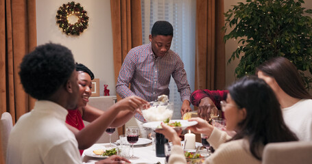 A young student of multi-ethnic background invited friends to a birthday party. The guy in a shirt is standing at the table and cutting meat. Friends pour themselves food and drink, talk, laugh, and