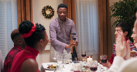 A multi-ethnic guy in a checkered shirt uncorks a bottle of wine with a corkscrew while sitting with friends at the festive table. The students are eating and chatting, and applaud as the guy smiles