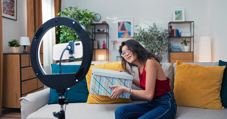 Beauty vlogger enthusiastically presents her clothing haul, opens a branded box with curiosity and joy, talks to followers about her fashion choices during a sponsored review.