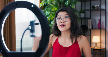 Asian beauty blogger is recording makeup tutorial video for watchers for channel, waving hand at phone set up on stand with a ring light, woman has cosmetics in front of her to review ads.