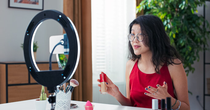 A popular influencer vlogger creates a video on social media talking into phone set up on ring light stand, a woman has facial care products in front of her, looks into the camera and smiles broadly. - Powered by Adobe