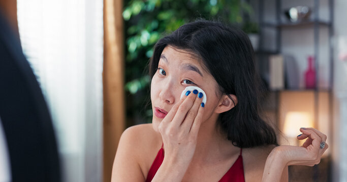 Female content creator films skincare vlog while cleansing face with white cotton pad. She looks in the mirror and records tutorial using ring light and phone from home.