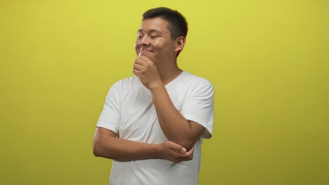 Hispanic young man in white t shirt with hand on chin and folded arm in yellow studio; thoughtful reflection.