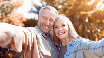 A joyful older couple smiles broadly while taking a selfie in a vibrant garden filled with colorful foliage. The sunlight creates a warm atmosphere as they enjoy their time together.