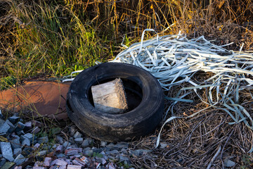A worn-out tire sits on the ground, holding a wooden block, surrounded by debris and tall grass during the golden hour of sunset